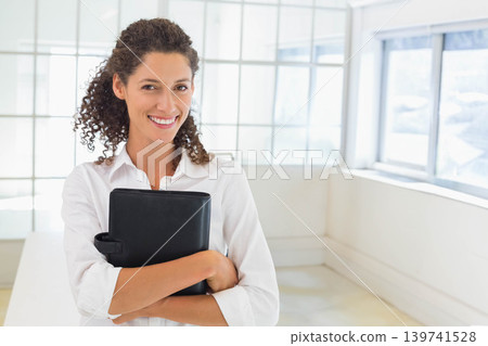 Adult woman standing holding black portfolio in bright office with grid windows wearing white shirt Adult woman standing holding black portfolio in bright office with grid windows wearing white shirt 139741528