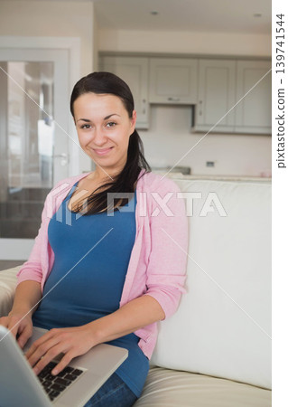 Adult woman sitting on couch in blue-top pink-cardigan typing on laptop at home, kitchen glass-door Adult woman sitting on couch in blue-top pink-cardigan typing on laptop at home, kitchen glass-door 139741544