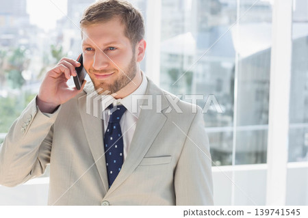 Man in light-gray suit, navy tie talking on smartphone by office windows overlooking skyline 139741545