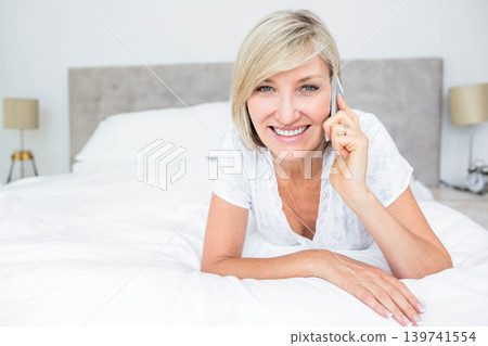 Mature woman reclining on white bedding in bedroom, wearing lace top and holding smartphone Mature woman reclining on white bedding in bedroom, wearing lace top and holding smartphone 139741554
