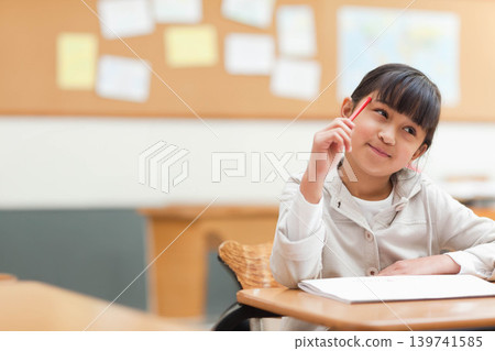 Asian child sitting in classroom at desk holding red pencil, open notebook with cork board visible Asian child sitting in classroom at desk holding red pencil, open notebook with cork board visible 139741585