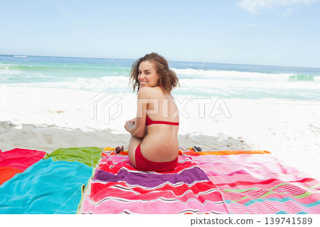 Woman lounging on striped towels at shoreline, wearing red swimsuit, sandals and sunglasses nearby Woman lounging on striped towels at shoreline, wearing red swimsuit, sandals and sunglasses nearby 139741589