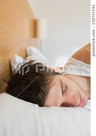 Man lying right side on bed, wearing white tank top, resting head on pillow near headboard Man lying right side on bed, wearing white tank top, resting head on pillow near headboard 139741591