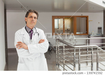 Midlife male doctor standing with crossed arms in hospital in lab coat with stethoscope, copy space Midlife male doctor standing with crossed arms in hospital in lab coat with stethoscope, copy space 139741592
