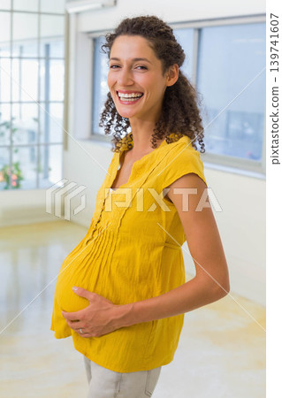 Pregnant woman standing in sunlit studio cradling bump wearing mustard top light pants wedding ring 139741607
