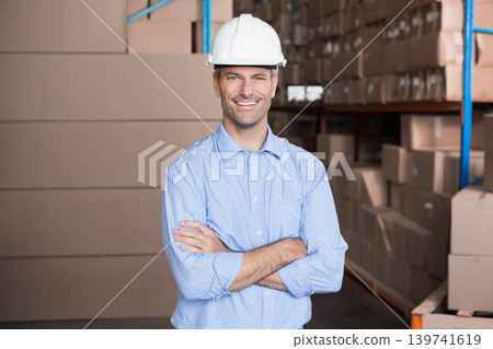 Man standing with arms crossed in warehouse among stacked boxes, wearing white hard hat 139741619