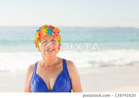 Senior woman standing, posing on sandy shore in blue swimsuit, floral cap, ocean waves horizon Senior woman standing, posing on sandy shore in blue swimsuit, floral cap, ocean waves horizon 139741620