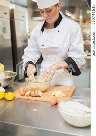 Female chef mixing batter in glass bowl at stainless counter with eggs, wearing chef coat, hat Female chef mixing batter in glass bowl at stainless counter with eggs, wearing chef coat, hat 139741630