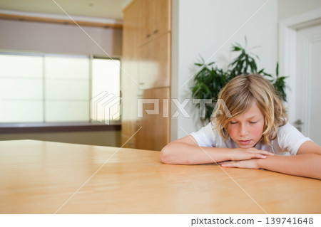 Boy leaning on large light-wood table at home, wearing white t-shirt, resting head, copy space Boy leaning on large light-wood table at home, wearing white t-shirt, resting head, copy space 139741648