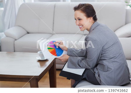 Woman in blazer sitting on sofa in lounge holding pie chart pointing with pen, copy space Woman in blazer sitting on sofa in lounge holding pie chart pointing with pen, copy space 139741662