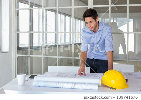 Man wearing blue shirt leaning over office table reviewing plans with yellow hard hat, copy space Man wearing blue shirt leaning over office table reviewing plans with yellow hard hat, copy space 139741670