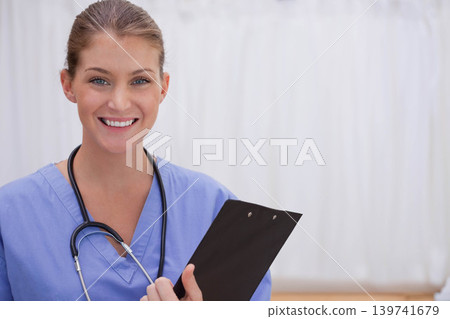 Female nurse posing by privacy drape in clinic in blue scrubs stethoscope holding clipboard smiling Female nurse posing by privacy drape in clinic in blue scrubs stethoscope holding clipboard smiling 139741679