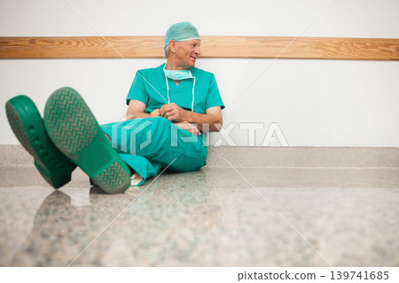 Senior male surgeon sitting in hospital hall leaning on wall by rail wearing scrubs mask clogs Senior male surgeon sitting in hospital hall leaning on wall by rail wearing scrubs mask clogs 139741685
