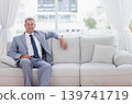 Middle-aged man sitting on pale gray sofa by large window wearing suit and wristwatch 139741719