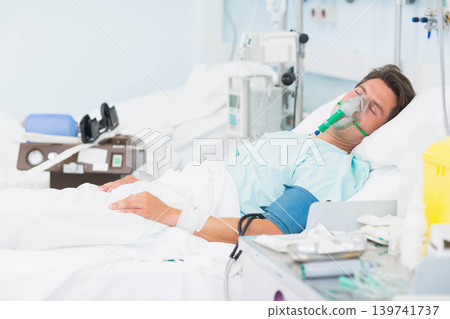 Middle-aged male resting in hospital bed wearing blue gown with oxygen mask, IV pump and cuff Middle-aged male resting in hospital bed wearing blue gown with oxygen mask, IV pump and cuff 139741737