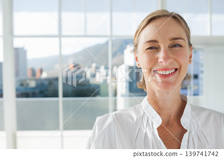 Mature woman smiling near windows with skyline, wearing white blouse and gold studs, copy space Mature woman smiling near windows with skyline, wearing white blouse and gold studs, copy space 139741742