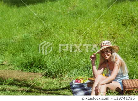 Woman sitting on checked blanket grassy slope holding straw hat and fruit basket beside, copy space Woman sitting on checked blanket grassy slope holding straw hat and fruit basket beside, copy space 139741786