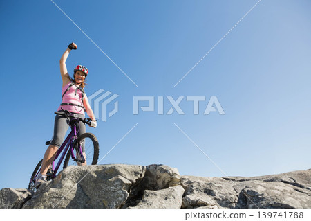 Mid-adult woman helmeted in gear cheering while standing on boulder with purple bike, copy space Mid-adult woman helmeted in gear cheering while standing on boulder with purple bike, copy space 139741788