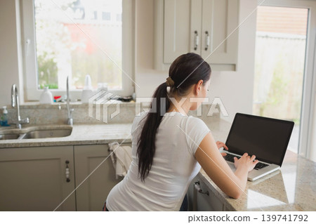 Woman leaning against granite kitchen counter typing on open laptop near sink faucet Woman leaning against granite kitchen counter typing on open laptop near sink faucet 139741792