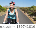 Female cyclist riding rural road holding handlebars, wearing helmet and hydration pack 139741810