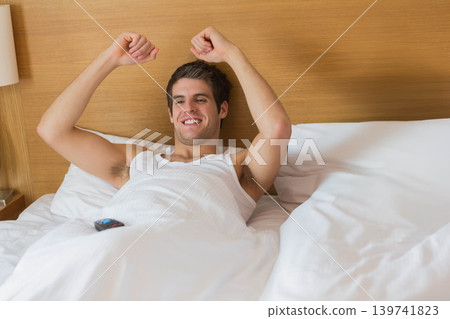 Man in his twenties reclining on bed stretching arms upward with clenched fists, remote on duvet 139741823