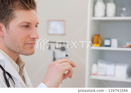 Male doctor wearing white coat preparing syringe in clinic with stethoscope and bottles, copy space Male doctor wearing white coat preparing syringe in clinic with stethoscope and bottles, copy space 139741846