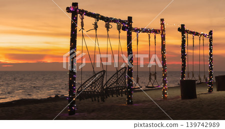 Swings on a beach are illuminated by lights as the sun sets over the sea. The sky shows warm colors while waves gently touch the shore. High quality photo Swings on a beach are illuminated by lights as the sun sets over the sea. The sky shows warm colors while waves gently touch the shore. High quality photo 139742989