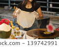 A woman prepares sweet roti at a Bangkok street stall, skillfully stretching the dough. She has gloves on her hands while oils and other ingredients are nearby. High quality photo 139742991