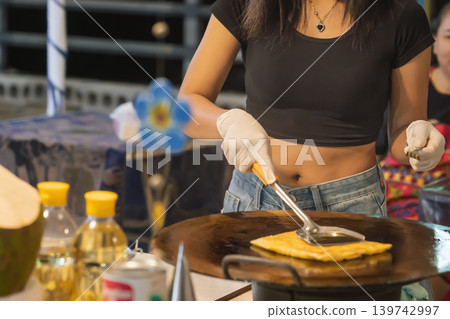 In Bangkok street market, a woman cooks roti on a hot surface while wearing gloves. She flips the dough with a spatula. The setting is lively with nearby vendors and customers. High quality photo In Bangkok street market, a woman cooks roti on a hot surface while wearing gloves. She flips the dough with a spatula. The setting is lively with nearby vendors and customers. High quality photo 139742997