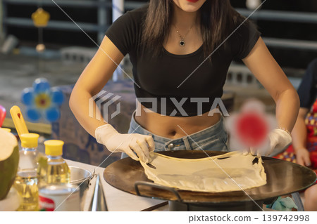 In a busy Bangkok street stall, a woman cooks a sweet roti pancake. She skillfully spreads the dough while customers watch and wait for their tasty treat. High quality photo 139742998
