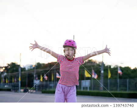 Happy little girl wearing a pink safety helmet raising her arms and smiling while roller skating outdoors. Happy little girl wearing a pink safety helmet raising her arms and smiling while roller skating outdoors. 139743740