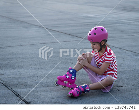 Little girl wearing a pink safety helmet crying and looking at her injured knee after falling down while skating. Little girl wearing a pink safety helmet crying and looking at her injured knee after falling down while skating. 139743741