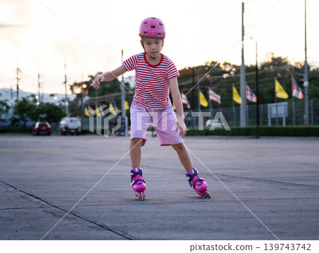 Little girl wearing a pink safety helmet practicing balance and roller skating at an outdoor plaza. Little girl wearing a pink safety helmet practicing balance and roller skating at an outdoor plaza. 139743742