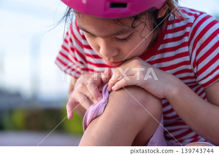 Little girl wearing a pink safety helmet crying and looking at her injured knee after falling down while skating. Little girl wearing a pink safety helmet crying and looking at her injured knee after falling down while skating. 139743744