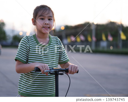 Happy little girl riding a kick scooter and smiling outdoors at a park during sunset. Happy little girl riding a kick scooter and smiling outdoors at a park during sunset. 139743745