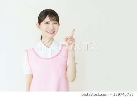 A portrait of a young Japanese female caregiver wearing a pink apron against a white background. 139743755