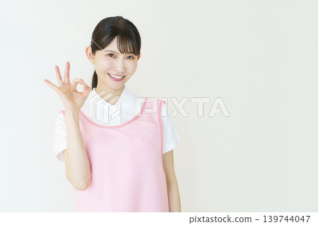 A portrait of a young Japanese female caregiver wearing a pink apron against a white background. 139744047