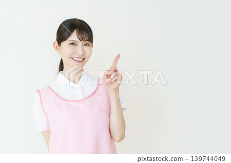 A portrait of a young Japanese female caregiver wearing a pink apron against a white background. A portrait of a young Japanese female caregiver wearing a pink apron against a white background. 139744049