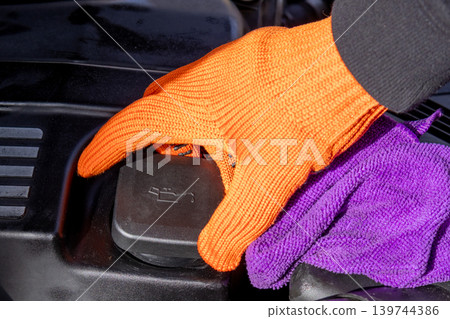 A mechanic wearing an orange glove checks the oil level in a car engine using a purple cloth in a garage setting 139744386