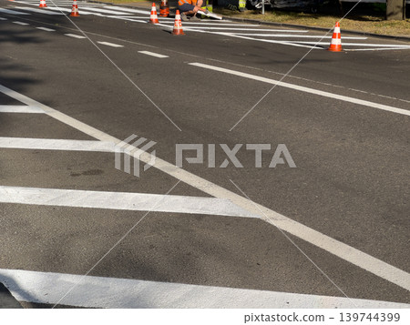 Workers are painting white road markings on a street. Safety cones are placed to guide traffic during the work Workers are painting white road markings on a street. Safety cones are placed to guide traffic during the work 139744399