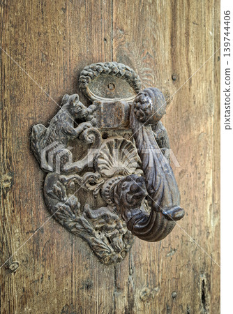A vintage cast iron door knocker features detailed animal and face carvings. It is mounted on a rustic wooden door that shows signs of age and weathering. A vintage cast iron door knocker features detailed animal and face carvings. It is mounted on a rustic wooden door that shows signs of age and weathering. 139744406