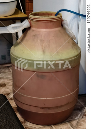 A large container sits in a kitchen with a hose attached. It appears to hold liquid for use in cooking or storage. Other kitchen items are present nearby 139744901