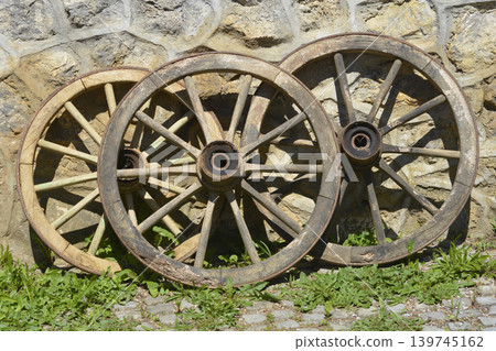 Three old wooden cart wheels leaning against a stone wall Three old wooden cart wheels leaning against a stone wall 139745162