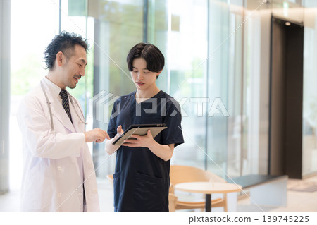 畫面中,醫生一邊看著平板電腦一邊給予指示和傳遞訊息,旁邊還有一位新來的醫生或護士,這體現了醫療領域的數位轉型(DX)和團隊合作。 畫面中,醫生一邊看著平板電腦一邊給予指示和傳遞訊息,旁邊還有一位新來的醫生或護士,這體現了醫療領域的數位轉型(DX)和團隊合作。 139745225
