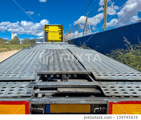 The shot shows the empty metal ramp and platform of a yellow car carrier truck parked beside a blue wall. Sunlight shines on the scene with a few clouds in the sky. The shot shows the empty metal ramp and platform of a yellow car carrier truck parked beside a blue wall. Sunlight shines on the scene with a few clouds in the sky. 139745819