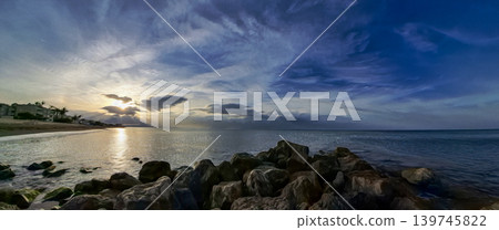 A wide scene shows a sunset over the sea. The sun sets on the horizon and reflects on the water. A rocky breakwater is in the foreground. Clouds fill the sky with interesting shapes. A wide scene shows a sunset over the sea. The sun sets on the horizon and reflects on the water. A rocky breakwater is in the foreground. Clouds fill the sky with interesting shapes. 139745822