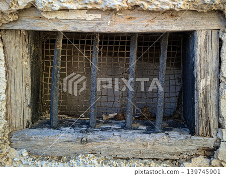 A small wooden window shows signs of age with rough textures. The window has an iron security grate and wire mesh, placed in a stucco wall. The setting looks historic and worn. A small wooden window shows signs of age with rough textures. The window has an iron security grate and wire mesh, placed in a stucco wall. The setting looks historic and worn. 139745901