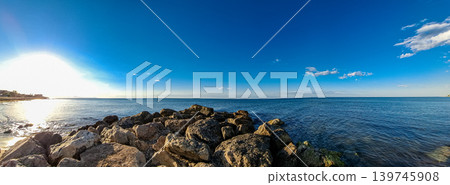 A rocky breakwater pier stretches into the Mediterranean Sea. The sun sets in the background, and the sky is bright blue. Calm waters reflect the warm light of the sun. A rocky breakwater pier stretches into the Mediterranean Sea. The sun sets in the background, and the sky is bright blue. Calm waters reflect the warm light of the sun. 139745908