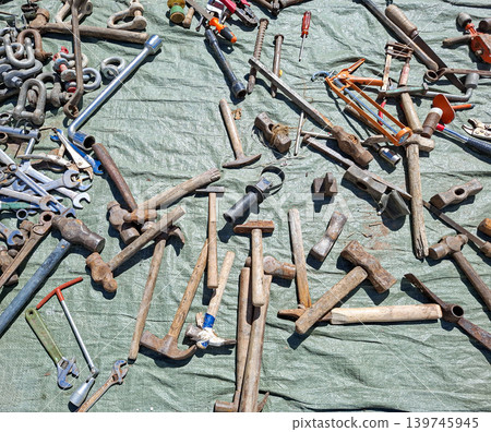 Various rusty hand tools are scattered on a wrinkled green tarp at an outdoor flea market. Old hammers, wrenches, screwdrivers, and pliers are visible among the items. Various rusty hand tools are scattered on a wrinkled green tarp at an outdoor flea market. Old hammers, wrenches, screwdrivers, and pliers are visible among the items. 139745945