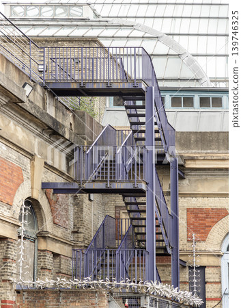 At Alexandra Palace, a deep purple-blue industrial staircase zigzags up a weathered brick and stone facade, its vertical railings creating a rhythmic geometric pattern. Below, a harsh line of razor At Alexandra Palace, a deep purple-blue industrial staircase zigzags up a weathered brick and stone facade, its vertical railings creating a rhythmic geometric pattern. Below, a harsh line of razor 139746325